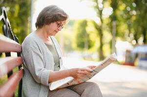 elegant ouderen vrouw in de overhemd is zittend Aan de bank in een park Aan een warm dag foto