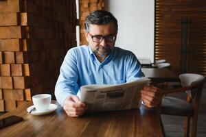 detailopname portret van echt senior knap Mens lezing krant, hebben koffie breken en zittend Bij tafel. foto