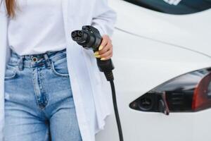 een meisjes hand- houdt een kabel voor opladen een elektrisch auto Aan de achtergrond van een wit auto. foto