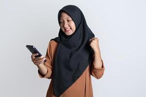 opgewonden mooi Aziatisch vrouw in bruin overhemd en hijab gebruik makend van mobiel telefoon, vieren succes, krijgen mooi zo nieuws geïsoleerd Aan wit achtergrond foto