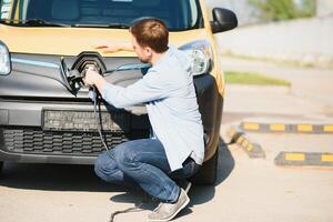 jong knap Mens Holding opladen kabel Bij elektrisch opladen station punt staand in de buurt zijn nieuw auto foto