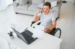 midden- leeftijd vrouw gebruik makend van laptop zittend Aan rolstoel Bij huis foto