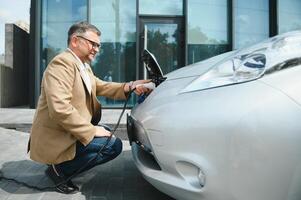 knap gebaard Mens zittend in de buurt zijn nieuw modern elektrisch auto en Holding plug van de oplader, terwijl auto is opladen Bij de opladen station foto