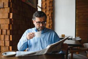 detailopname portret van echt senior knap Mens lezing krant, hebben koffie breken en zittend Bij tafel. foto