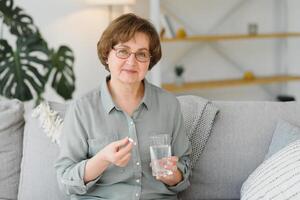 volwassen senior midden- oud vrouw Holding pil en glas van water nemen pijnstiller naar verlichten pijn, geneeskunde supplementen vitamines, antibiotica medicatie, medicijnen voor oud persoon concept, dichtbij omhoog visie. foto