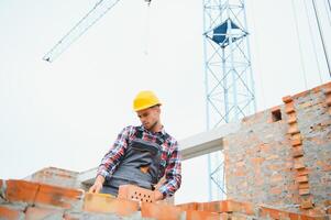 gebruik makend van bakstenen. jong bouw arbeider in uniform is bezig Bij de onvoltooid gebouw foto