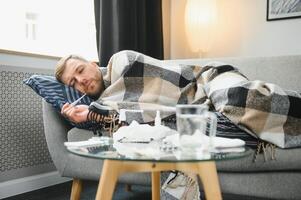 ziek gebaard Mens wie heeft slecht verkoudheid of seizoensgebonden griep zittend Aan bankstel Bij huis. vent met koorts vervelend warm plaid rillingen met bezorgd gezicht uitdrukking. foto