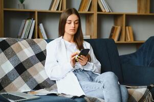 jong gelukkig Kaukasisch vrouw ze Jaren 20 in gewoontjes kleren houden in hand- gebruik mobiel cel telefoon zitten Aan blauw sofa binnen- rust uit Bij huis in eigen kamer appartement. mensen levensstijl concept foto