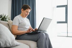 technologie, mensen en communicatie concept - gelukkig senior vrouw met tablet pc computer hebben video babbelen Bij huis. modern middelbare leeftijd Jaren 50 grootmoeder zitten kom tot rust Aan bankstel in leven kamer gebruik makend van laptop. foto