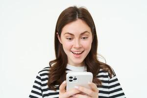 portret van jong vrouw op zoek Bij smartphone scherm met verrast, versteld staan gezicht uitdrukking, lezing Super goed nieuws Aan telefoon foto