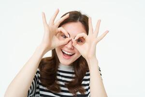portret van Vrolijk, mooi jong vrouw, maakt bril met vingers in de omgeving van ogen en lachend, hebben plezier, genieten van evenement, staand over- wit achtergrond foto