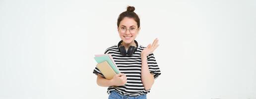 portret van meisje in bril, met hoofdtelefoons over- haar nek, houdt notebooks en studie materiaal voor Universiteit of middelbare school, looks gelukkig en verrast, wit achtergrond foto