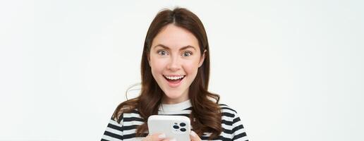 portret van opgewonden brunette meisje, reageren naar verbazingwekkend promo Aan smartphone, Holding mobiel telefoon en lachend, geïsoleerd Aan wit achtergrond foto