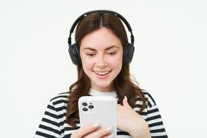 beeld van jong vrouw in koptelefoon, gebruik makend van smartphone en lachend, aan het kijken video Aan mobiel telefoon, luistert naar muziek- Aan streaming onderhoud app, wit achtergrond foto