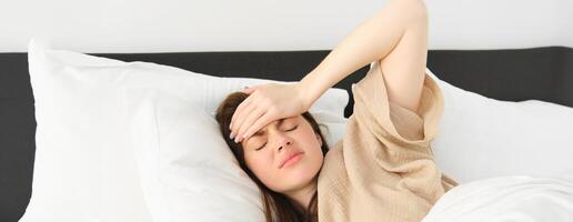 vrouw aan het liegen in bed en gevoel onwel, hebben hoofdpijn, aanraken haar hoofd met gefrustreerd gezicht, heeft hoog koorts of migraine. concept van Gezondheid en mensen foto