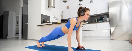 portret van vrouw in draadloze koptelefoon, aan het doen training van huis, staand in plank met sport- apparatuur, luisteren muziek, op zoek zelfverzekerd foto