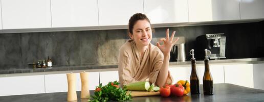 portret van glimlachen brunette meisje maken veganistisch diner, tonen Oke teken, aanbevelen gezond voedsel, hakken groenten in keuken voor salade of voor veganistisch ontbijt foto