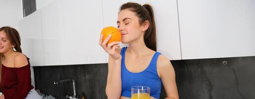 portret van geschiktheid instructeur, glimlachen vrouw, geniet geur van vers fruit, drinken oranje sap en op zoek Vrolijk, staand in keuken in activewear foto