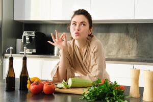 portret van mooi jong vrouw in badjas, Koken maaltijd voor familie, shows koks kus, Oke teken, maken voedsel, voorbereidingen treffen vegetarisch diner, hakken groenten foto