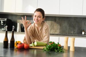 portret van glimlachen brunette meisje maken veganistisch diner, tonen Oke teken, aanbevelen gezond voedsel, hakken groenten in keuken voor salade of voor veganistisch ontbijt foto