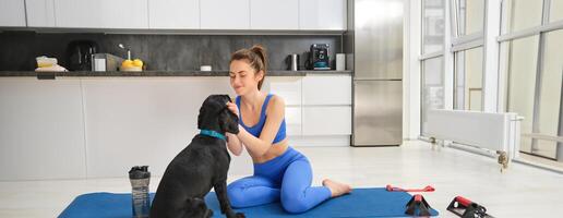 beeld van jong vrouw doet training van huis, zit Aan yoga mat in leven kamer en Toneelstukken met haar zwart hond, puppy leidt af meisje van aan het doen opdrachten foto