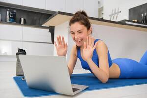 portret van mooi, fit jong vrouw Aan verdieping Bij huis, gezegde Hallo Bij laptop scherm, golvend hand- en lachend, training van een afstand met vriend, aan het doen yoga klasse online foto