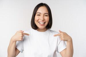gelukkig en zelfverzekerd Aziatisch vrouw, leerling glimlachen en richten Bij haarzelf, zelfpromotie, tonen logo Aan t-shirt, staand over- wit achtergrond foto