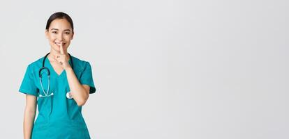 covid19, gezondheidszorg arbeiders, pandemisch concept. vrolijk, glimlachen vrouw Aziatisch verpleegster in scrubs hebben geheim, maken verrassing, tonen zwijgen gebaar, shhh houden stil, staand wit achtergrond foto