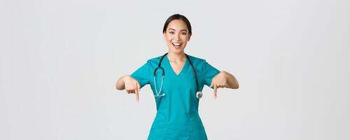 covid19, gezondheidszorg arbeiders, pandemisch concept. gelukkig glimlachen mooi Aziatisch verpleegster, vrouw dokter in scrubs richten omlaag, uitnodigend voor controle, tonen ziekenhuis advertentie, wit achtergrond foto