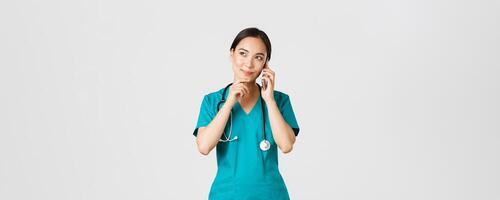 covid19, gezondheidszorg arbeiders en voorkomen virus concept. portret van glimlachen Aziatisch vrouw dokter, intern in scrubs pratend Aan telefoon en op zoek attent, denken of maken keuze foto