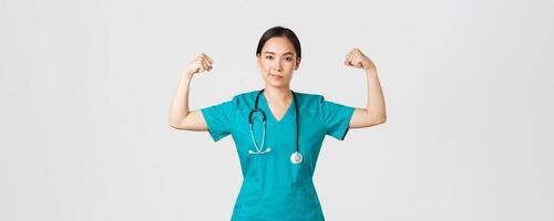 covid19, gezondheidszorg arbeiders en voorkomen virus concept. zelfverzekerd professioneel Aziatisch vrouw dokter, verpleegster in scrubs glimlachen brutaal en buigen biceps, wezen krachtig, Opschepper spieren foto