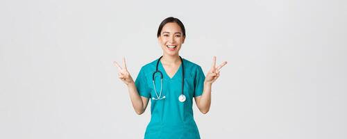 covid19, gezondheidszorg arbeiders, pandemisch concept. gelukkig Aziatisch vrouw dokter, verpleegster in scrubs tonen vrede kawaii gebaar en lachend, blijven positief gedurende werk in ziekenhuis, wit achtergrond foto