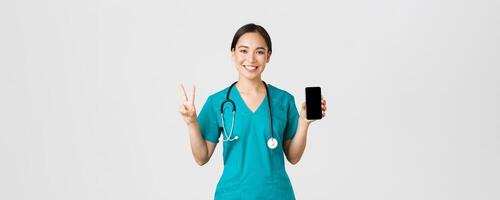 covid19, gezondheidszorg arbeiders en online geneeskunde concept. glimlachen zelfverzekerd Aziatisch vrouw dokter, arts in scrubs tonen vrede teken en mobiel scherm, promoten overleg app, wit achtergrond foto