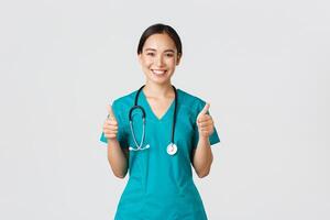 covid19, gezondheidszorg arbeiders, pandemisch concept. professioneel zelfverzekerd glimlachen dokter, vrouw arts in scrubs tonen duimen omhoog met verzekerd uitdrukking, ervoor zorgen allemaal goed, perfect of uitstekend foto