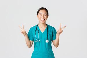 covid19, gezondheidszorg arbeiders, pandemisch concept. gelukkig Aziatisch vrouw dokter, verpleegster in scrubs tonen vrede kawaii gebaar en lachend, blijven positief gedurende werk in ziekenhuis, wit achtergrond foto