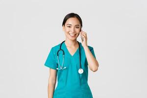 covid19, gezondheidszorg arbeiders en voorkomen virus concept. glimlachen gelukkig Aziatisch vrouw dokter, jong intern in scrubs pratend Aan telefoon, op zoek Bij camera hoopvol, wit achtergrond foto