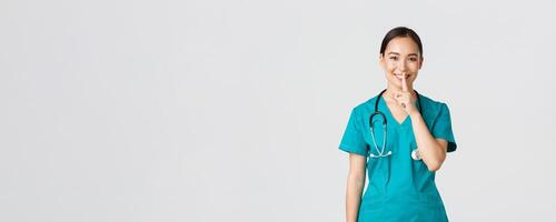 covid19, gezondheidszorg arbeiders, pandemisch concept. vrolijk, glimlachen vrouw Aziatisch verpleegster in scrubs hebben geheim, maken verrassing, tonen zwijgen gebaar, shhh houden stil, staand wit achtergrond foto