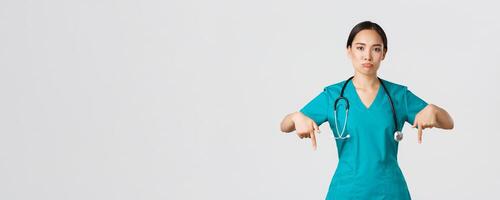 covid19, gezondheidszorg arbeiders, pandemisch concept. van streek en teleurgesteld pruilen Aziatisch vrouw therapeut, verpleegster in scrubs richten vingers omlaag, mokkend verdrietig, tonen iets met betreuren foto