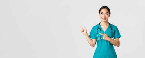 covid19, gezondheidszorg arbeiders, pandemisch concept. gefascineerd mooi Aziatisch vrouw intern, verpleegster in scrubs op zoek en richten bovenste links hoek met nieuwsgierig dromerig glimlach, wit achtergrond foto