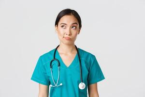 gezondheidszorg arbeiders, voorkomen virus, quarantaine campagne concept. attent Aziatisch vrouw dokter, verpleegster in scrubs op zoek bovenste links hoek en grijns onzeker, denken, wit achtergrond foto