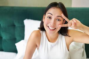 positief Aziatisch vrouw aan het liegen in bed, tonen vrede teken, geniet gelukkig ochtend, ontwaken omhoog vrolijk, blijven in haar slaapkamer foto