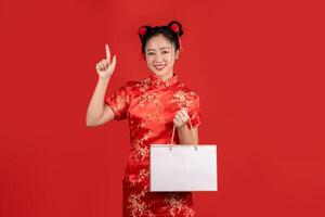 gelukkig Aziatisch vrouw vervelend rood qipao jurk Holding boodschappen doen zak geïsoleerd Aan rood achtergrond. gelukkig Chinese nieuw jaar foto