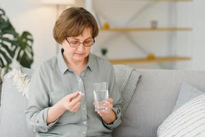 volwassen senior midden- oud vrouw Holding pil en glas van water nemen pijnstiller naar verlichten pijn, geneeskunde supplementen vitamines, antibiotica medicatie, medicijnen voor oud persoon concept, dichtbij omhoog visie. foto