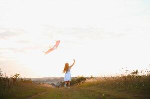 weinig schattig 7 jaren oud meisje rennen in de veld- met vlieger Aan zomer dag foto