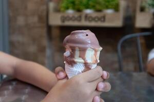 een jong kind hand- Holding een druipend chocola ijs room ijshoorntje in de cafe foto