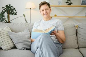 middelbare leeftijd huisvrouw zittend Aan sofa met boek, vrij tijd pensioen, hobby. foto