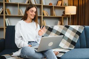 glimlachen aantrekkelijk jong vrouw zittend Aan sofa gebruik makend van laptop communiceren werken online Bij huis, gelukkig tiener meisje typen Aan computer, genieten van schrijven blog of chatten met vrienden in sociaal netwerk foto