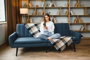 jong gelukkig Kaukasisch vrouw ze Jaren 20 in gewoontjes kleren houden in hand- gebruik mobiel cel telefoon zitten Aan blauw sofa binnen- rust uit Bij huis in eigen kamer appartement. mensen levensstijl concept foto