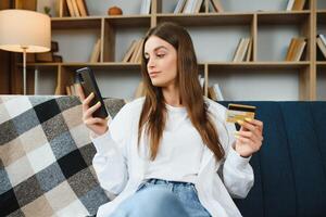 gelukkig tiener aan het doen boodschappen doen online zittend Aan bankstel Bij huis. jong vrouw insert credit kaart aantal Aan website met haar slim telefoon. foto