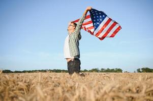 jong patriottisch boer staat tussen nieuw oogst. jongen wandelen met de Amerikaans vlag Aan de tarwe veld- vieren nationaal onafhankelijkheid dag. 4e van juli concept. foto